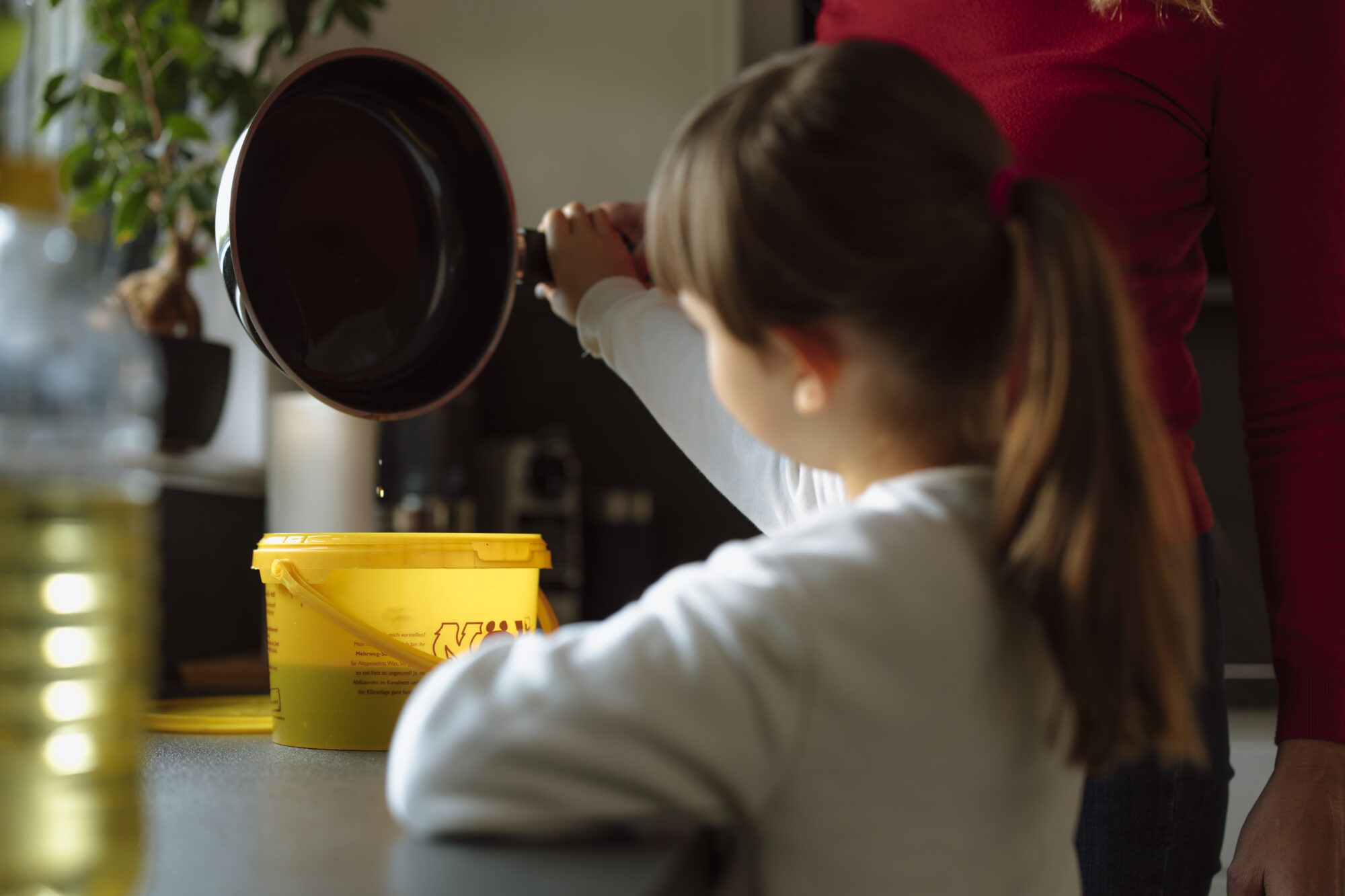 Auf dem Bild ist die Entsorgung von Altspeiseöl abgebildet. Ein Kind hält eine Pfanne mit Öl gemeinsam mit einer Frau in die Höhe. Öl tropft von der Pfanne in den gelben NÖLI Kübel. Im Vordergrund ist eine Ölflasche verschwommen zu erkennen.