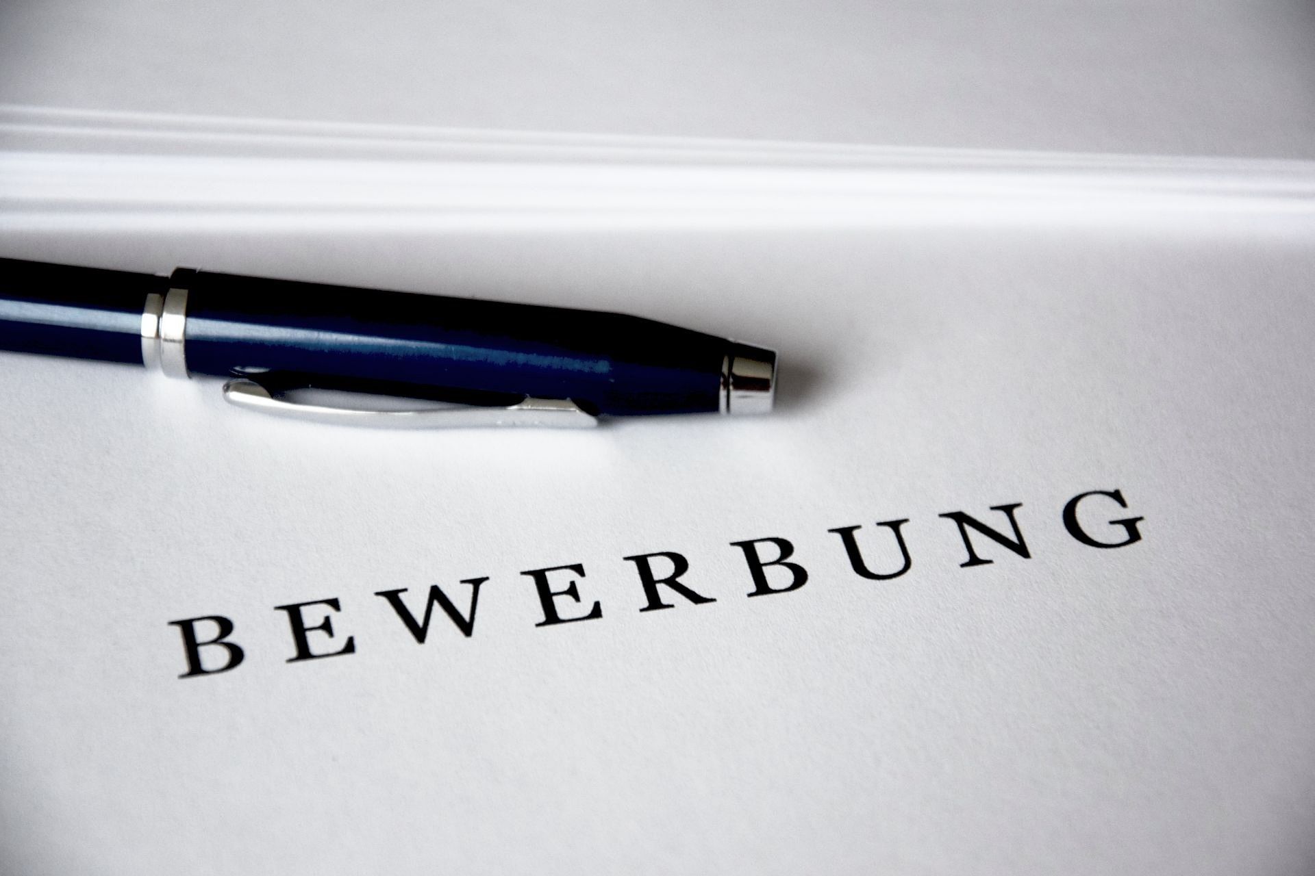 teilweise sichtbarer Schreibstift liegt auf weißem Papier, auf dem das Wort "Bewerbung" gedruckt ist