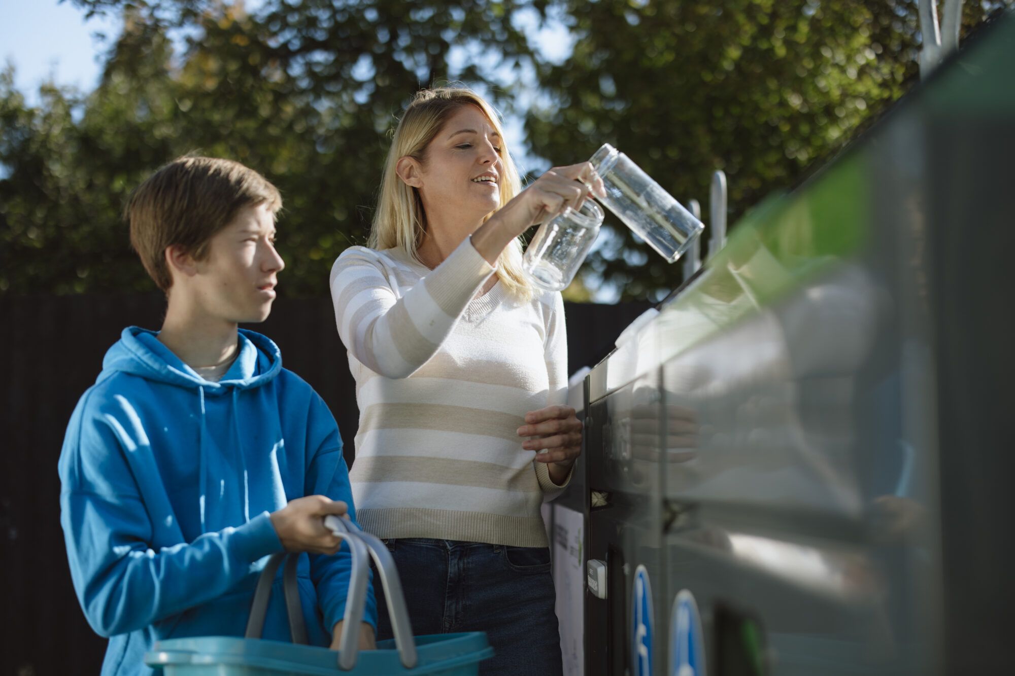 Eine Frau und ein Kind entsorgen Weißglas auf einer Sammelinsel. Der Bub mit braunen Haaren trägt einen blauen Pullover und hält einen blauen Korb in der Hand. Neben dem Bub steht eine Frau mit blonden Haaren und trägt einen Pullover und einer Jeans. Sie hält zwei weiße Konservengläser in der Hand und hält sie in Richtung Weißglas Container.