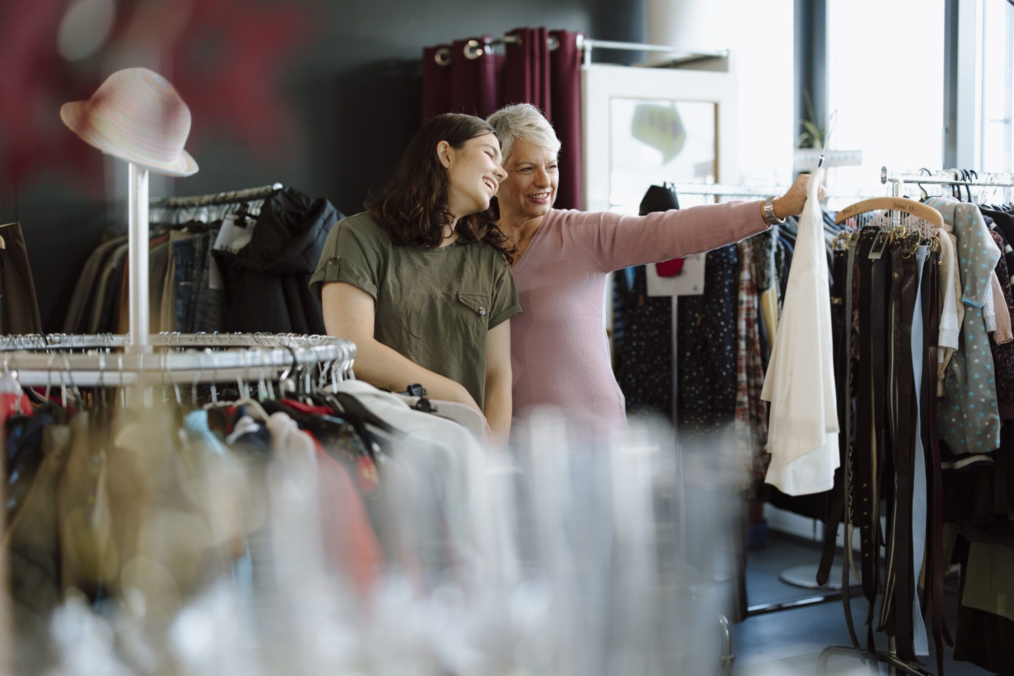 Ein jugendliches Mädchen und eine ältere Damen stehen in einem Secondhandshop in der Kleiderabteilung. Beide Frauen lächeln eine weiße Bluse an, welche die ältere Damen in der Hand hält. Die Jugendliche trägt leicht gewelltes braunes schulterlanges Haar und ein Kurzarm-T-Shirt in olive. Die ältere Dame hat kurzes grau-weißes Haar und trägt einen altrosafarbenen dünnen Strickpullover. Im Hintergrund im Bild sieht man Kleiderstangen mit unterschiedlichen Secondhand-Kleiderstücken wie Jeans, Westen, Gürtel und Babykleidung.