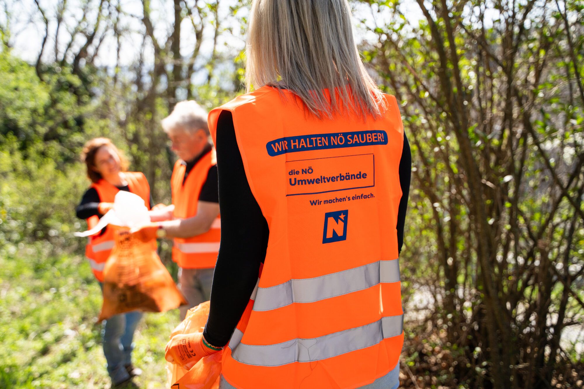 3 Personen, 2 im Hintergrund etwas unscharf, eine im Vordergrund mit dem R&uuml;cken zum Betrachter schauend. Alle tragen orange Warnwesten