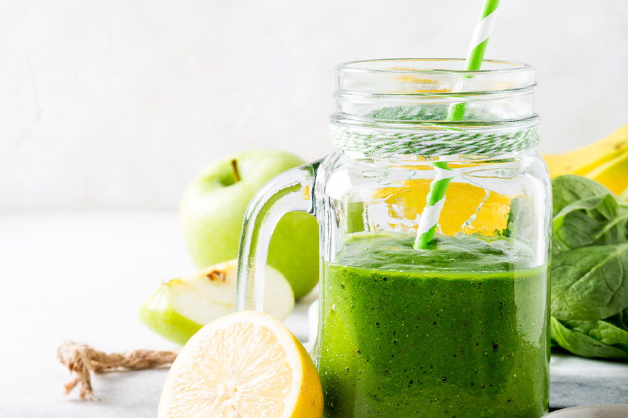 Ein Schraubglas mit Griff gefüllt mit leuchtend grünem Smoothie, ein grün-weiß gestreifter Strohhalm ragt heraus. Rundherum sind Spinatblätter, ein grüner Apfel und eine halbe Zitrone zu sehen.