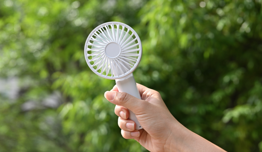 Zu sehen in eine Hand, die einen weißen Handventilator hält. Im Hintergrund ist Natur zu sehen.