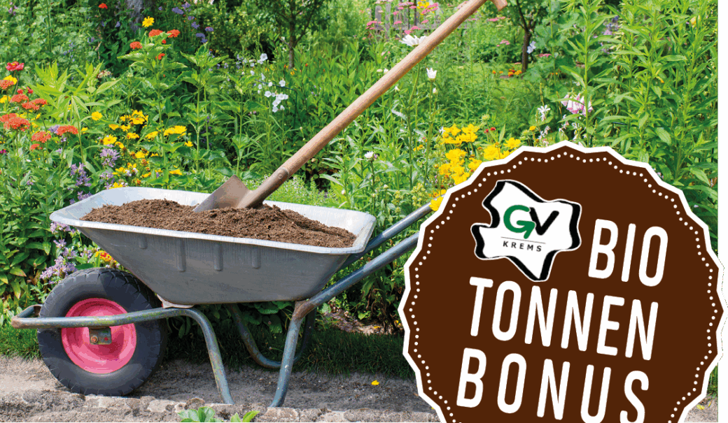 scheibtruhe mit Komposterde befüllt in der eine Schaufel steckt, steht vor grünem Beet im Garten, am linken Rand ist ein brauner Stempel mit dem GV Krems Biotonnenbonus
