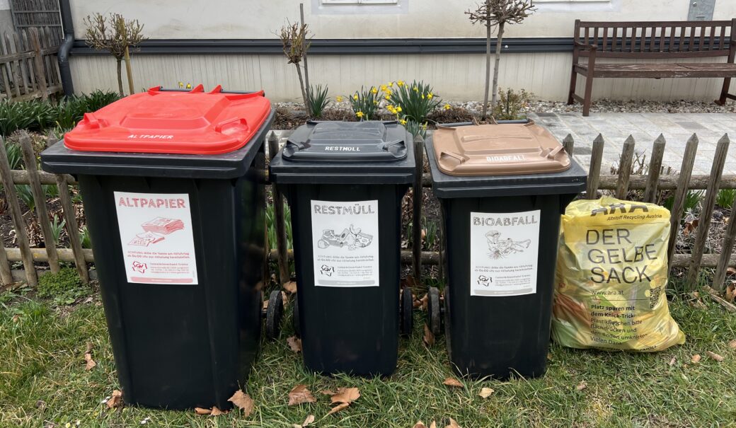 Altpapiertonne, Restmülltonne, Biomülltonne und Gelber Sack stehen vor dem Gartenzaun eines Einfamilienhauses.